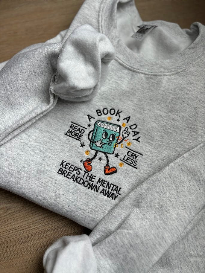 Embroidered sweatshirt with the phrase 'A Book a Day Keeps the Mental Breakdown Away' in bold font. Cozy and witty design for book lovers who find solace in reading. Perfect for anyone who values mental wellness and a good book. Multiple colors avail
