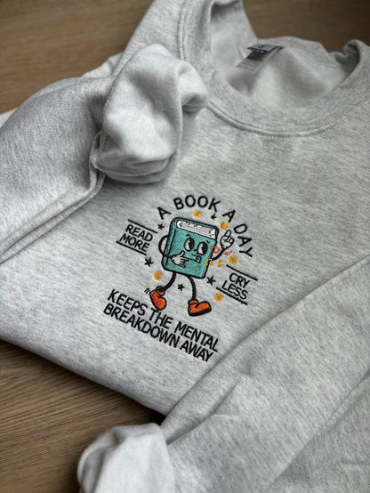 Embroidered sweatshirt with the phrase &#39;A Book a Day Keeps the Mental Breakdown Away&#39; in bold font. Cozy and witty design for book lovers who find solace in reading. Perfect for anyone who values mental wellness and a good book. Multiple colors avail