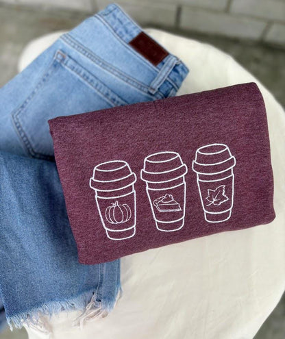 Heather Sport Dark Maroon Sweatshirt with 3 coffee cups on them. 1 cup has an outline of a pumpkin, 1 cup has an outline of a pie, the last cup has an outline of a leaf.