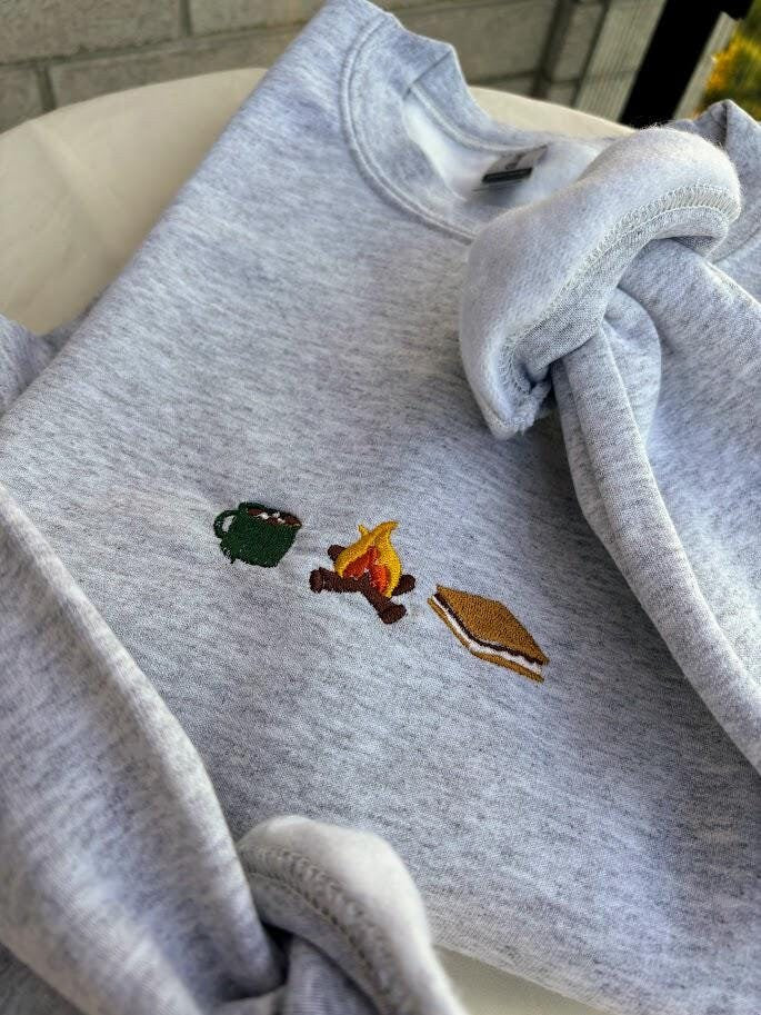 Embroidered sweatshirt featuring a cozy fall design with a steaming cup of hot chocolate, a glowing campfire, and delicious s&#39;mores. Perfect for autumn lovers and outdoor enthusiasts, this cozy apparel captures the warmth and joy of the season