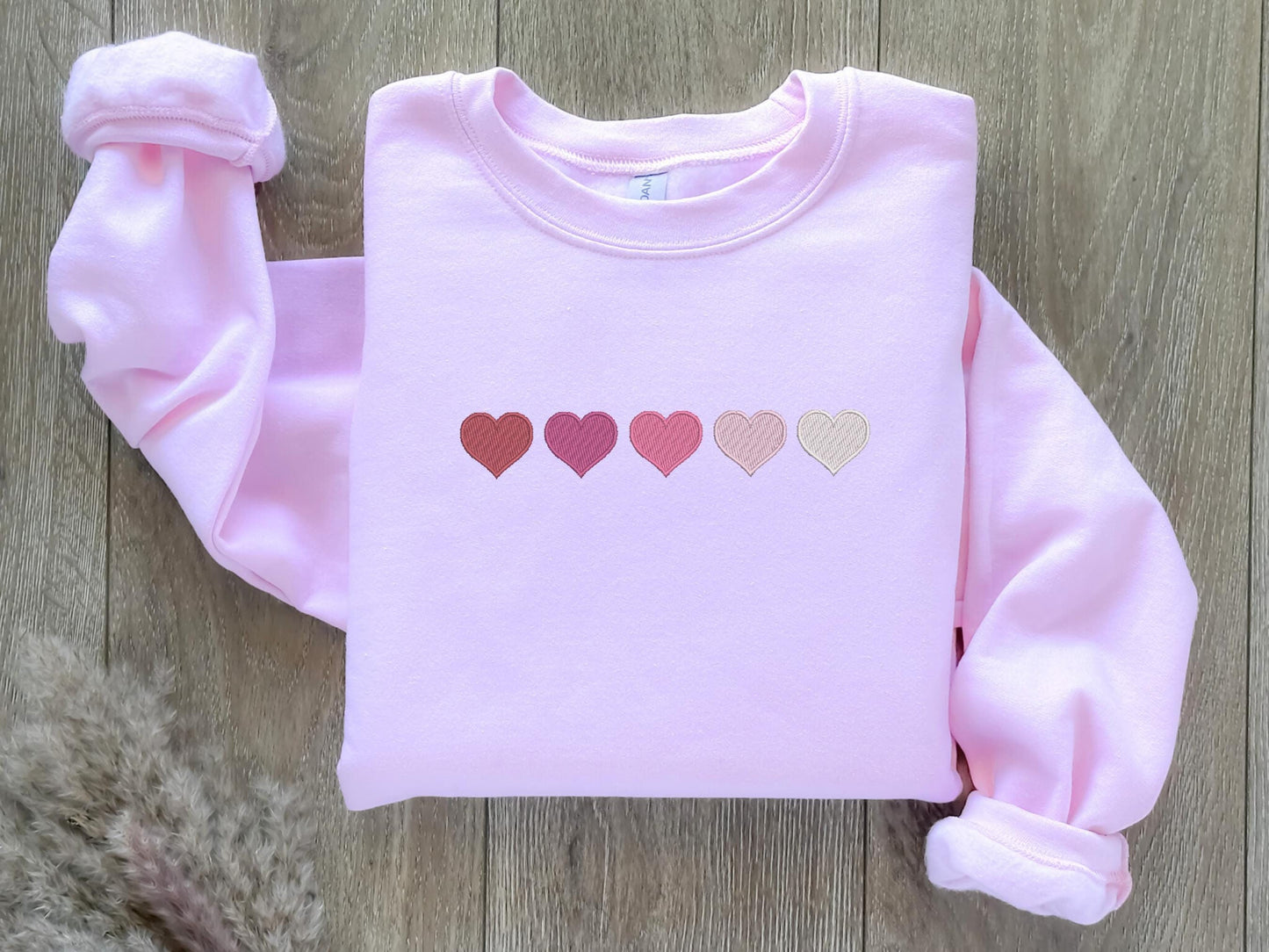 An embroidered sweatshirt featuring five hearts arranged in a row, transitioning from bold red to dark pink, slightly lighter pink, very light pink, and white. The gradient design adds a sweet and romantic touch, perfect for casual or cozy outfits.