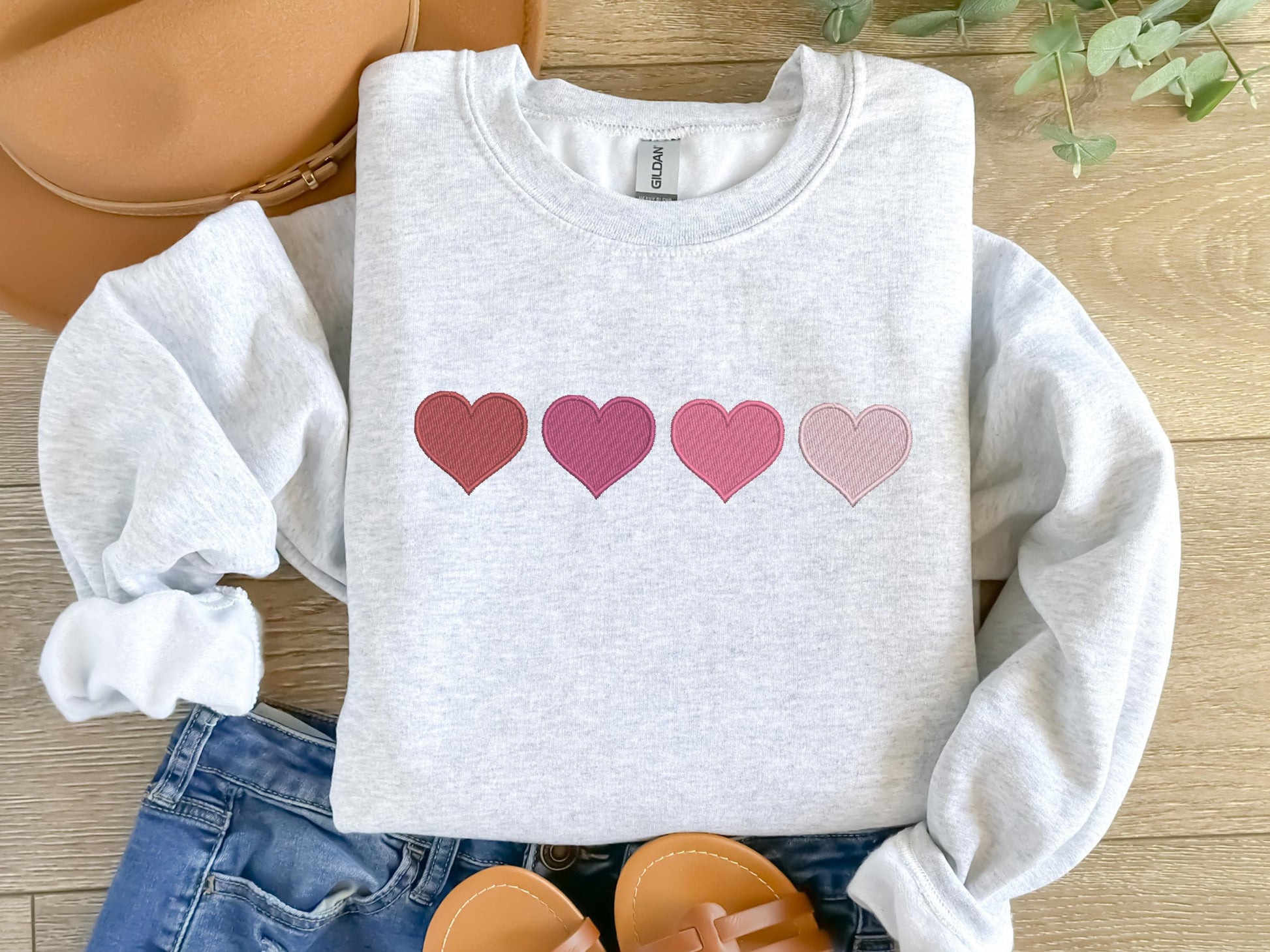 An embroidered sweatshirt featuring four hearts arranged in a row, each in a different shade of pink and one red. The design is minimal yet charming, perfect for a cozy and stylish look.