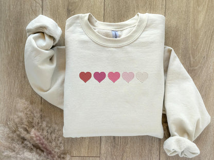 An embroidered sweatshirt featuring five hearts arranged in a row, transitioning from bold red to dark pink, slightly lighter pink, very light pink, and white. The gradient design adds a sweet and romantic touch, perfect for casual or cozy outfits