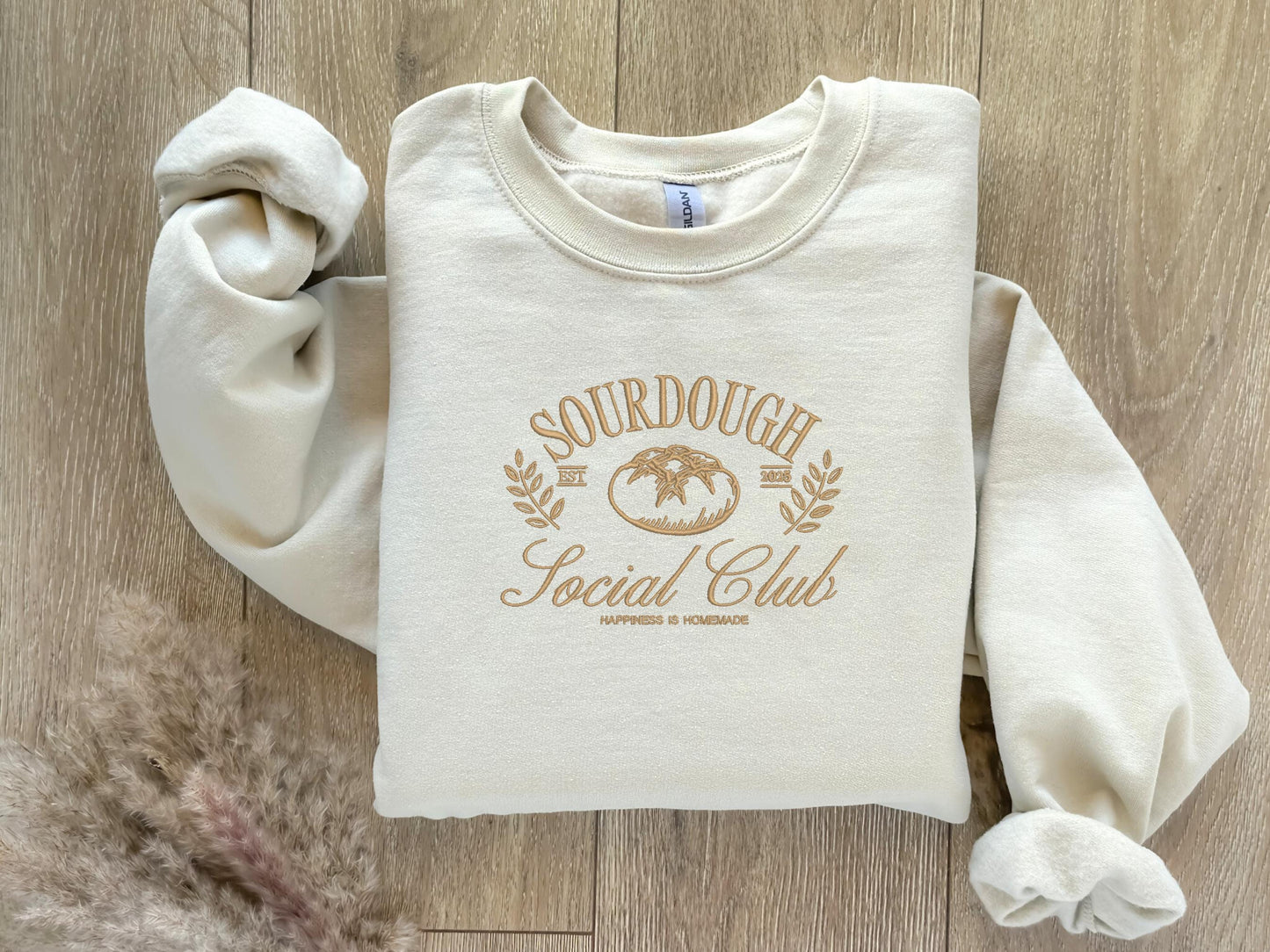 Beige-colored embroidered sweatshirt featuring the phrase &quot;Sourdough Social Club&quot; in a vintage-style font, arched around a detailed sourdough bread illustration in the center. Cozy and perfect for bread lovers and baking enthusiasts.